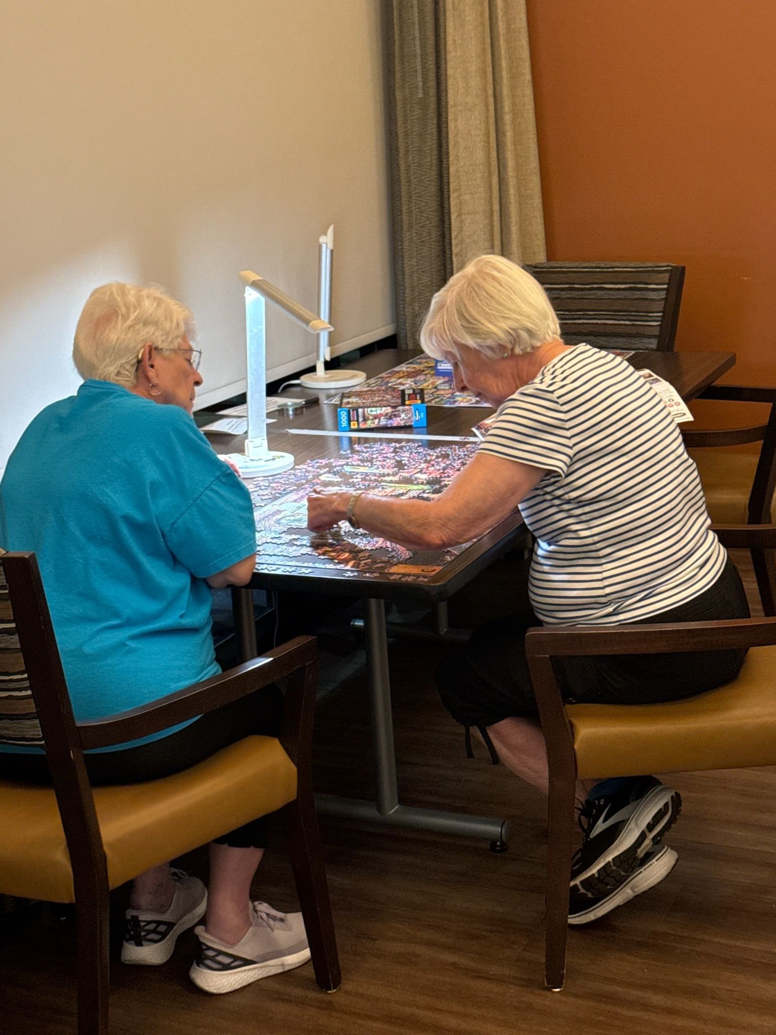 Two people seated, assembling a jigsaw puzzle at a table indoors. One wears blue, the other black and white stripes.