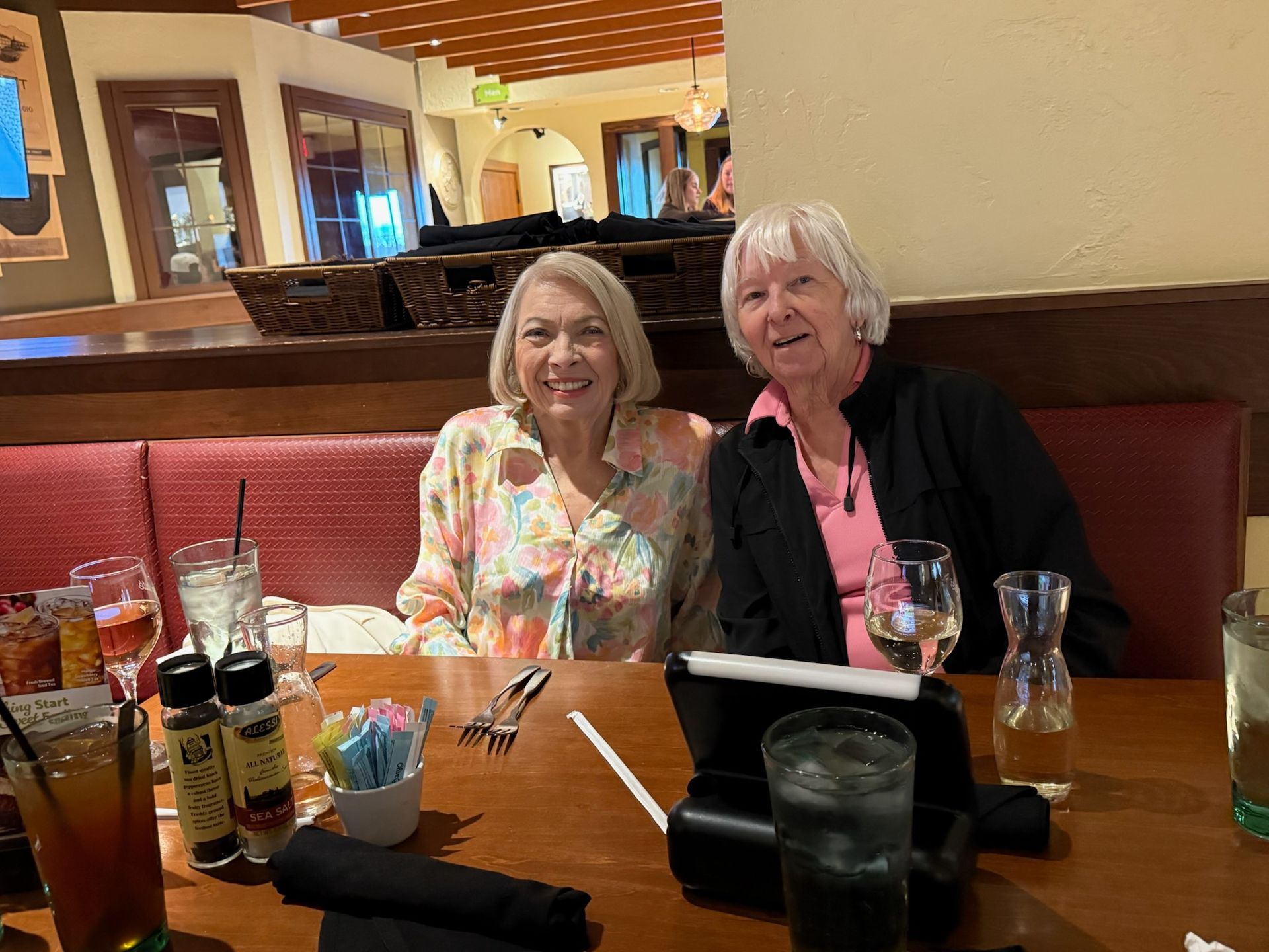 Two women seated at a restaurant table. One in a floral shirt, the other in a black jacket, smiling.