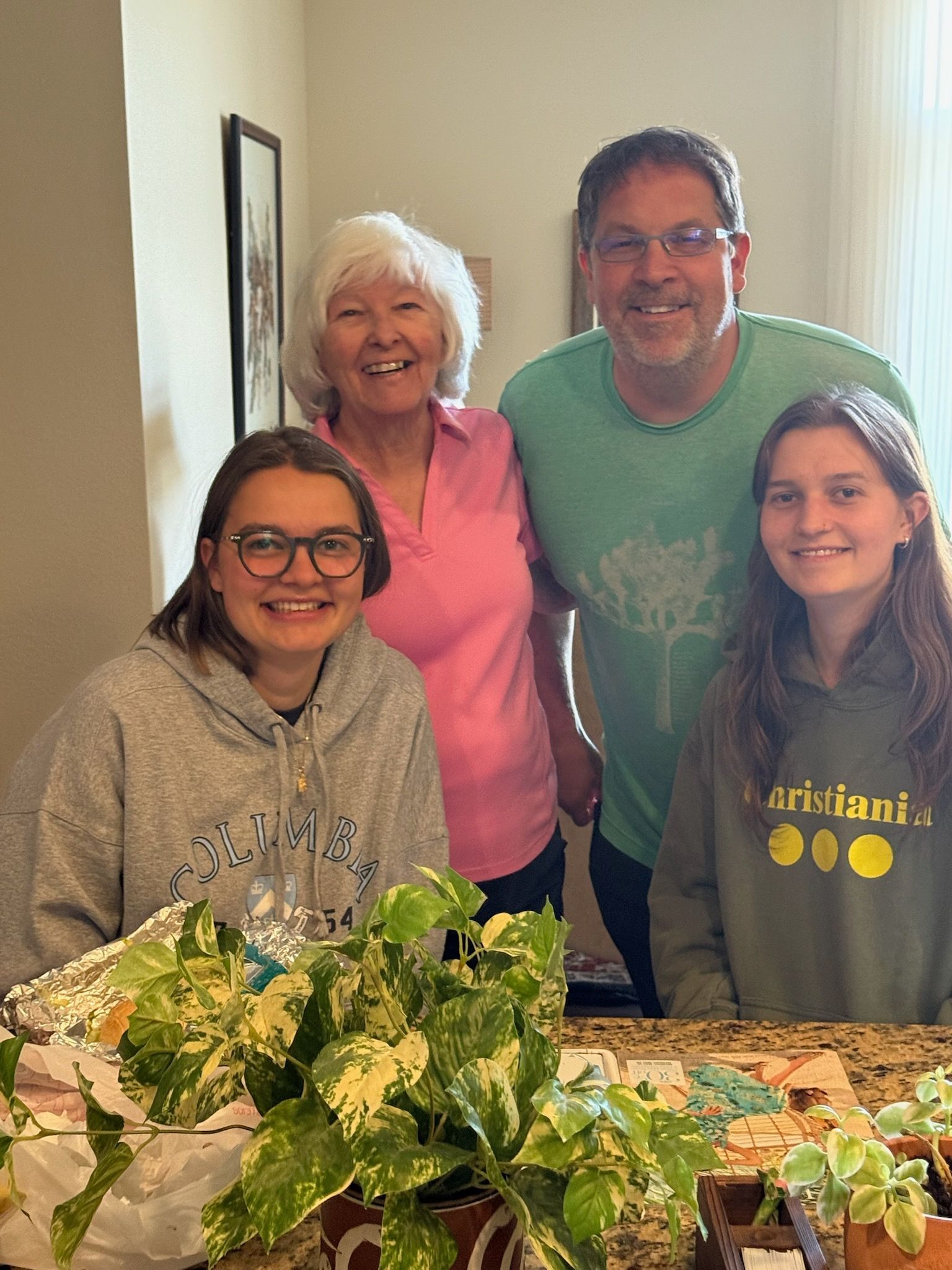 Family of four smiles together. An older woman in pink and a man in green stand behind two girls, one in a grey hoodie, one in green, in front of plants.