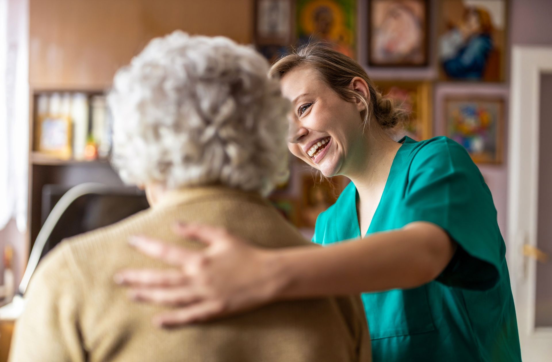 Caregiver smiles at an older person, placing a supportive hand on the person's back; indoors.