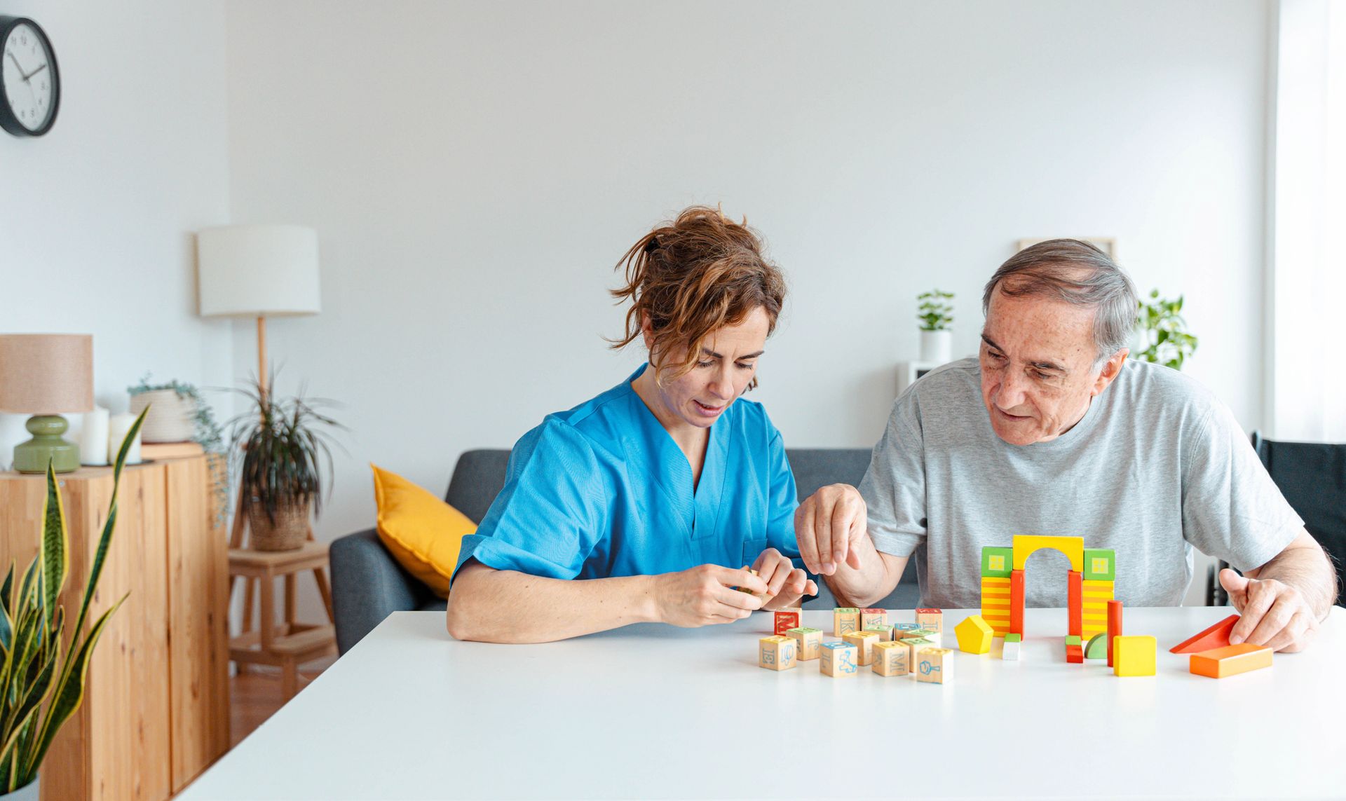 Woman in blue scrubs and older man building with colorful blocks at a table.