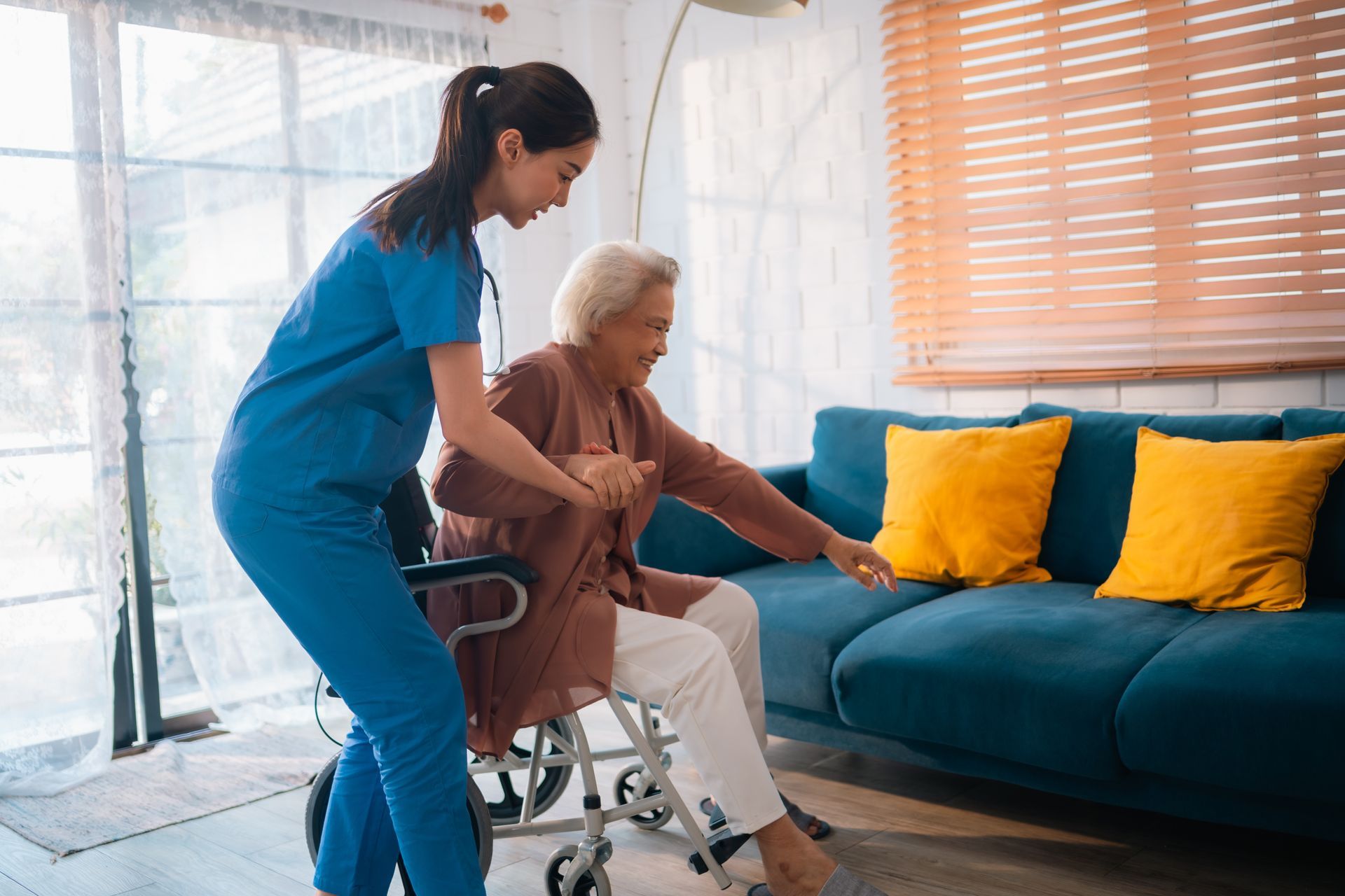 Caregiver in blue scrubs assisting an elderly person from a wheelchair to a couch.