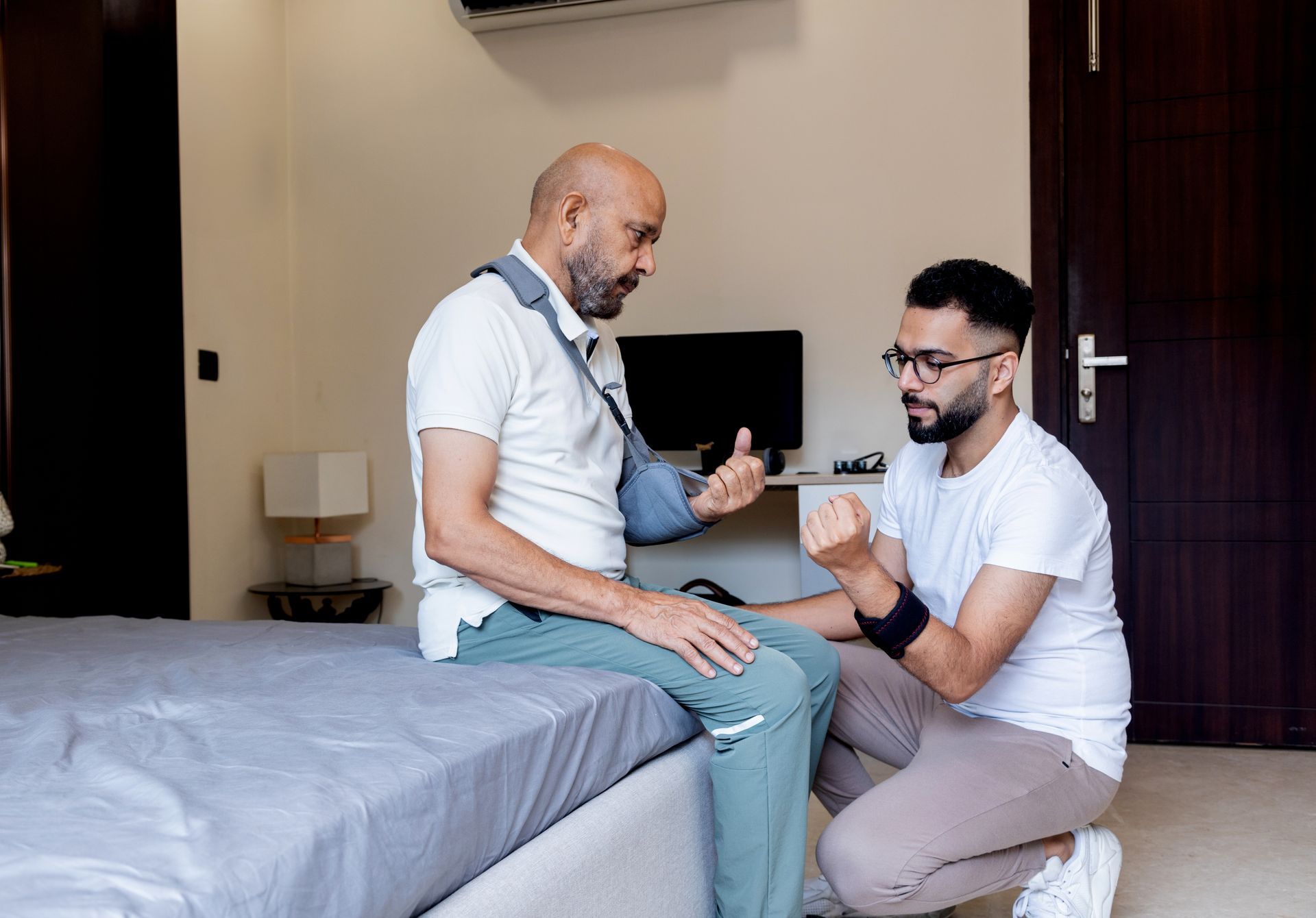 Man with arm brace sits on bed as another man kneels, assisting him, in a room.