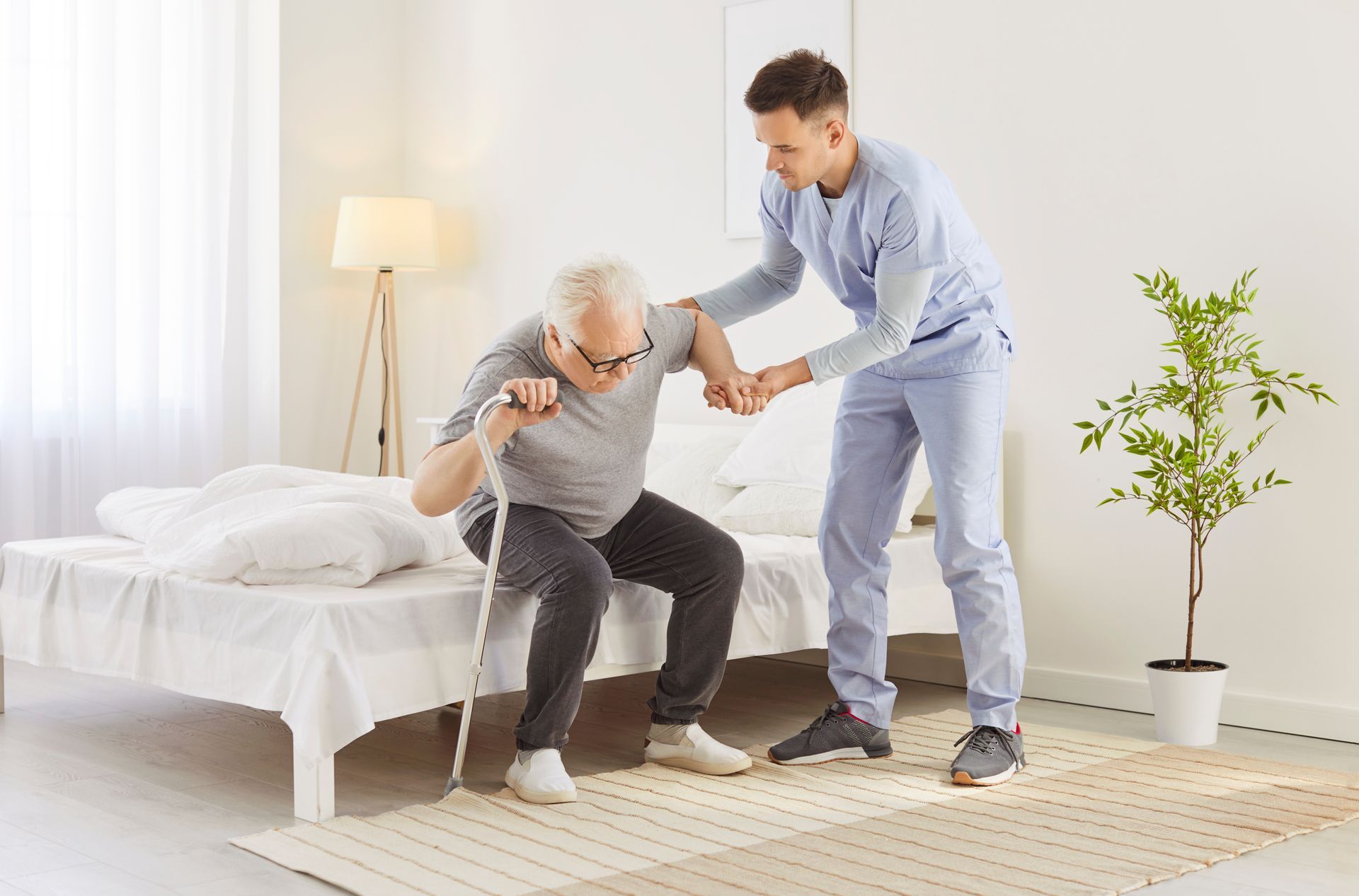 A caregiver helps an elderly person sit up from a bed, holding their arm for support.
