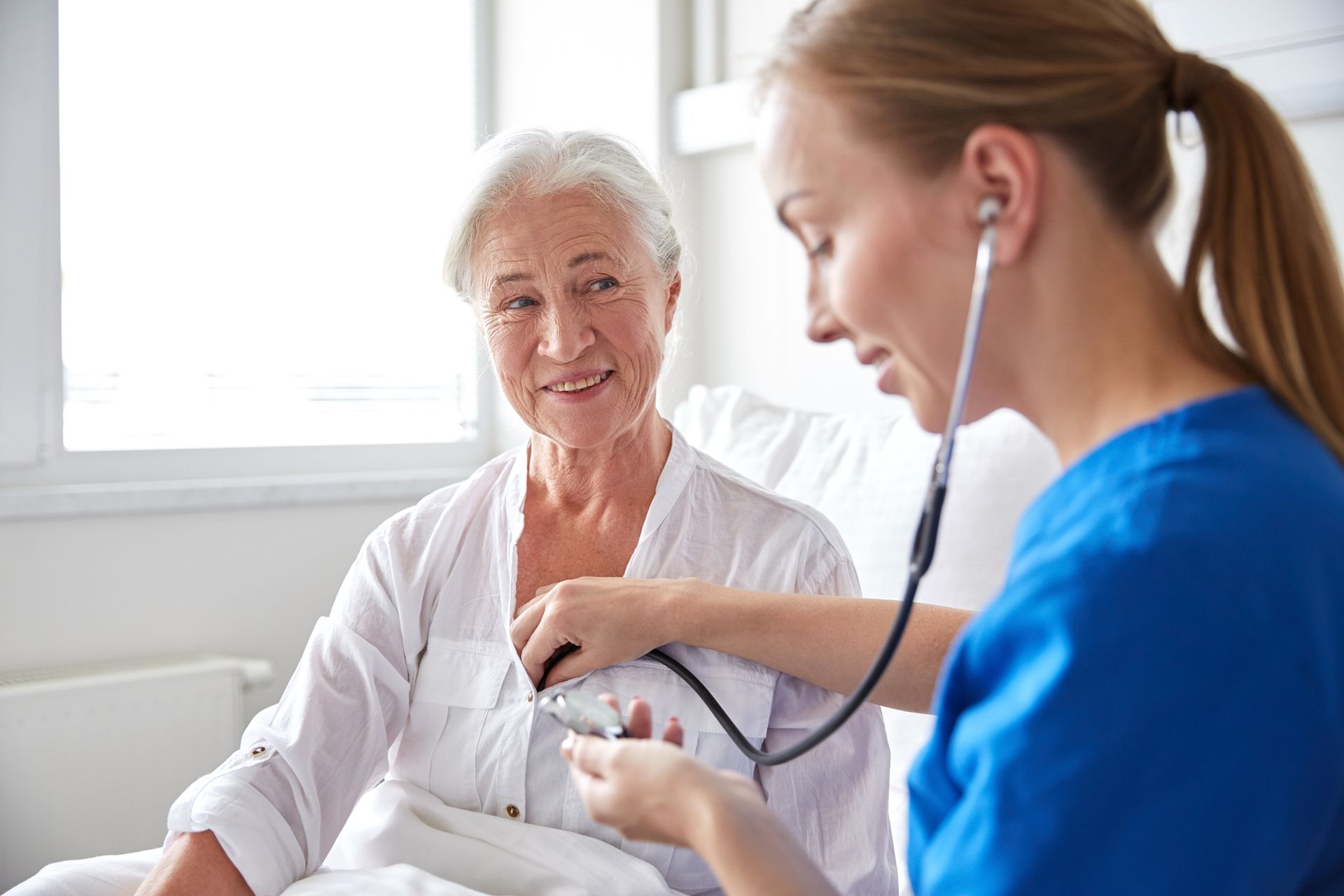 Nurse using a stethoscope to listen to an elderly person's chest in a light-filled room.