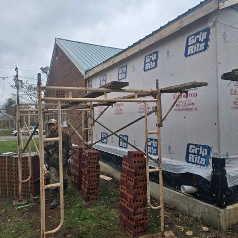 Man standing by bricks and scaffolding at a bricklaying construction site. Building covered in protective wrap.