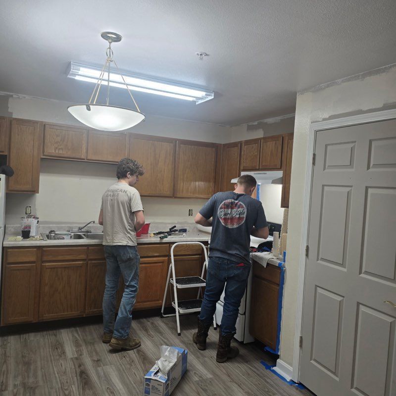 Two people painting a kitchen. One person stands at the sink, the other at the stove. Cabinets are wood-toned.
