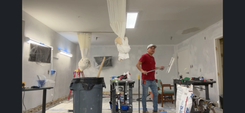 A man in a red shirt and jeans holds a tool in a room under construction.