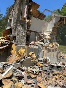 Damaged stone house, partially collapsed. Debris and exposed structure visible. Outdoor setting.