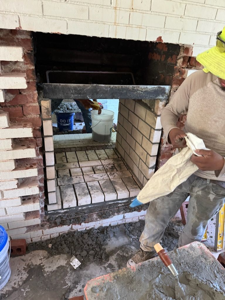 Construction worker laying bricks inside a fireplace. Red and white bricks, mortar, and tools visible.
