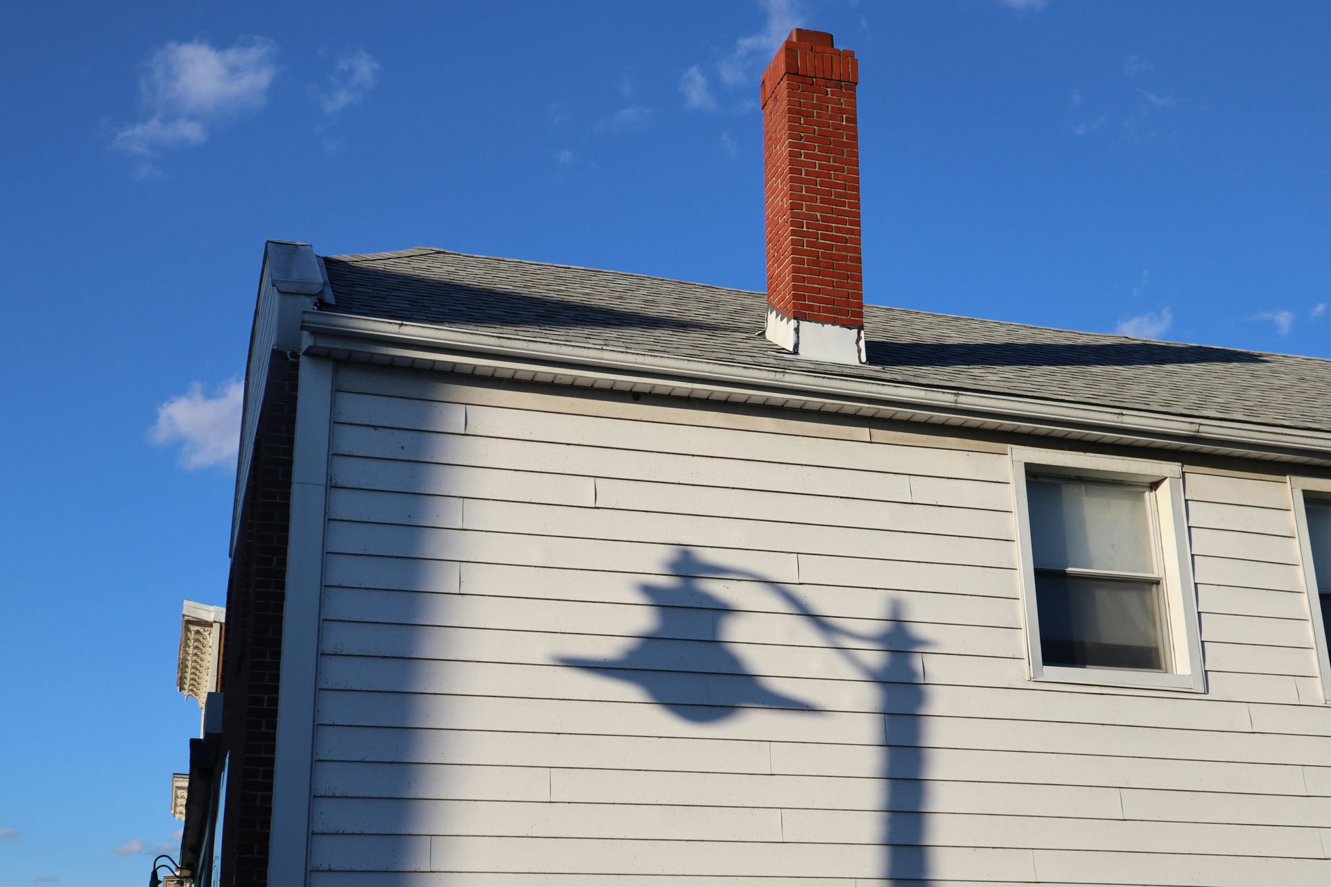 A building with a brick chimney and white siding, casting a shadow of a street lamp on the wall; blue sky.