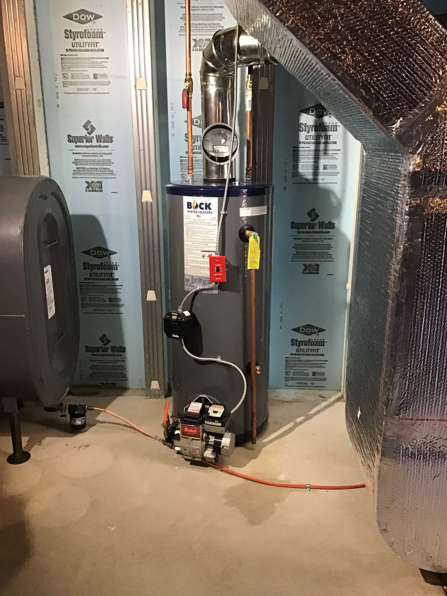 A water heater is sitting in a basement next to a fan.
