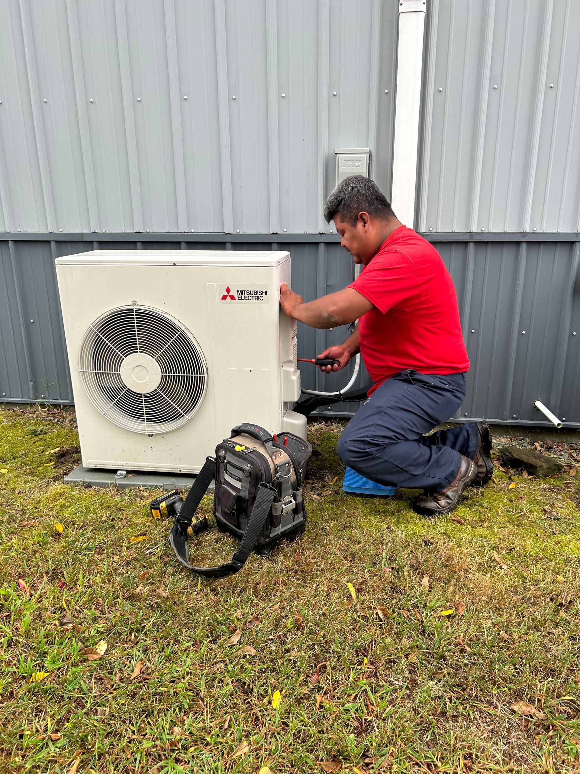 A man in a red shirt is kneeling down in front of an air conditioner.
