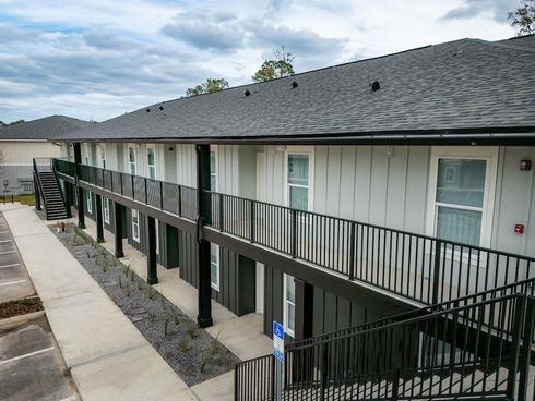 Two-story apartment building with dark gray roof, black railings, and light gray siding under a cloudy sky.