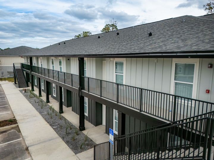Two-story apartment building with dark gray roof, black railings, and light gray siding under a cloudy sky.