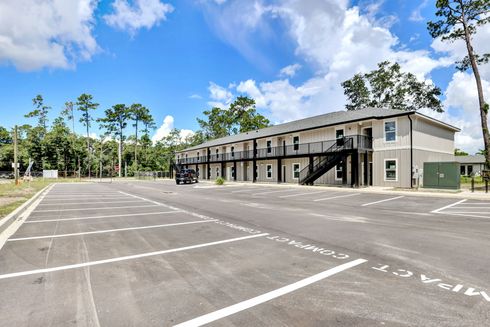 Two-story light gray building with black balconies and stairs, large empty parking lot.