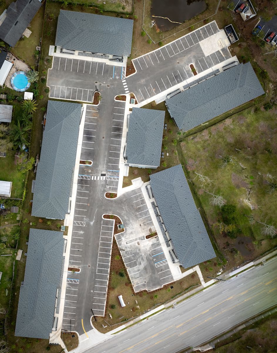 Aerial view of apartment complex with parking areas, asphalt driveways, and rectangular buildings with gray roofs.
