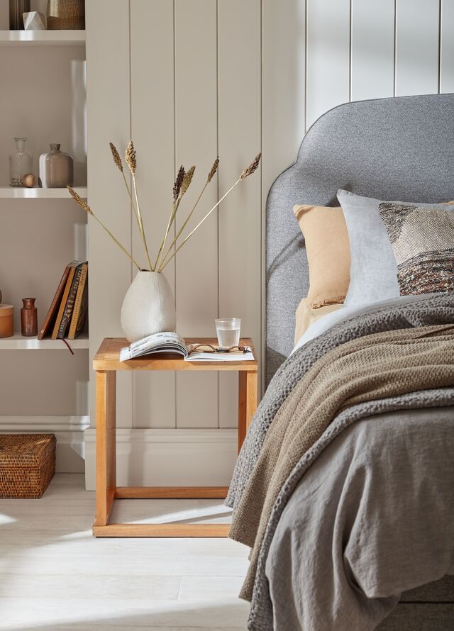 Cozy bedroom: gray bed with throw blanket, bedside table with vase and decor, wooden shelves with books and items.