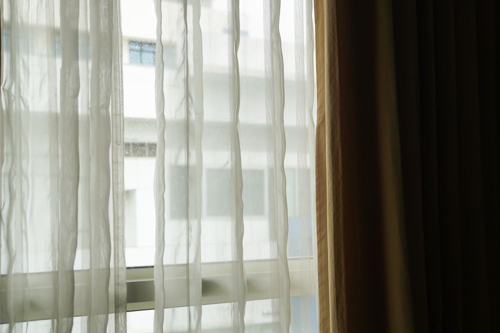 A Close Up of a Window With White Curtains and a Building in the Background — Tully Blinds in Cardwell, QLD