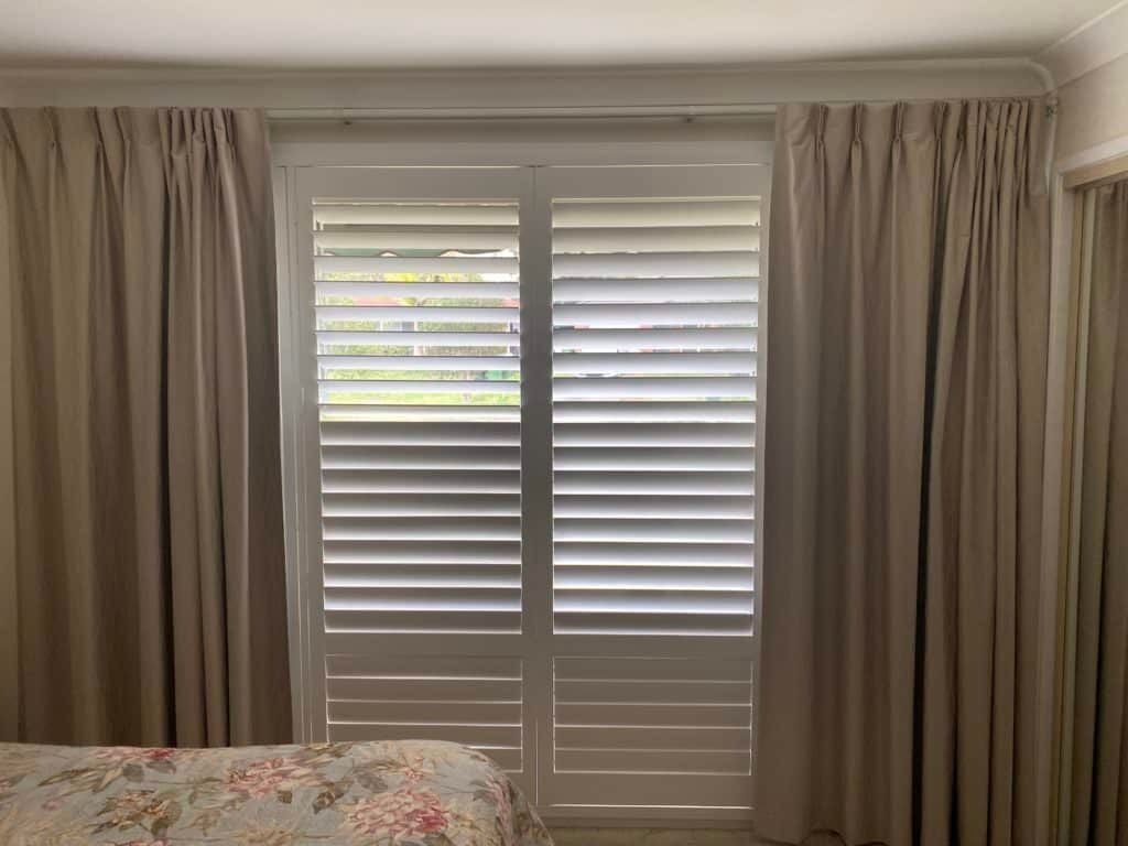 A Bedroom With a Bed, Curtains and Shutters on the Window — Tully Blinds in Tully, QLD