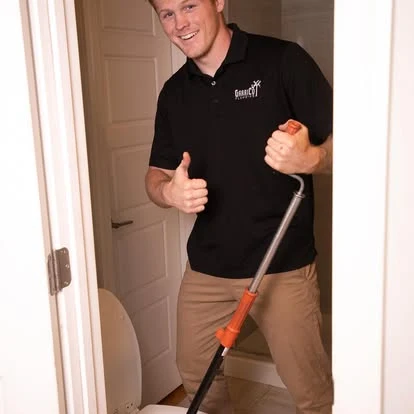 A person in a black polo shirt giving a thumbs up while using an orange and metal toilet auger in a bathroom.