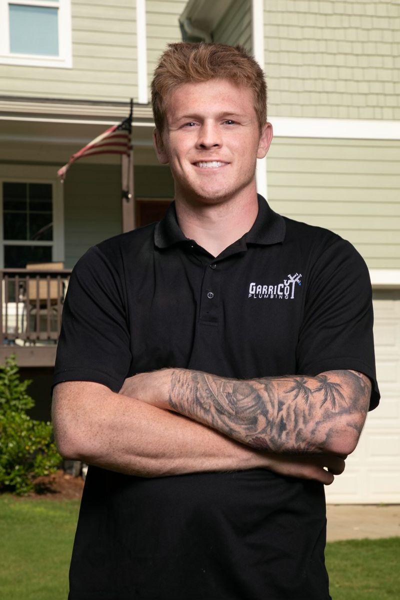A person wearing a black polo shirt with a company logo and tattooed arms stands with arms crossed outdoors.