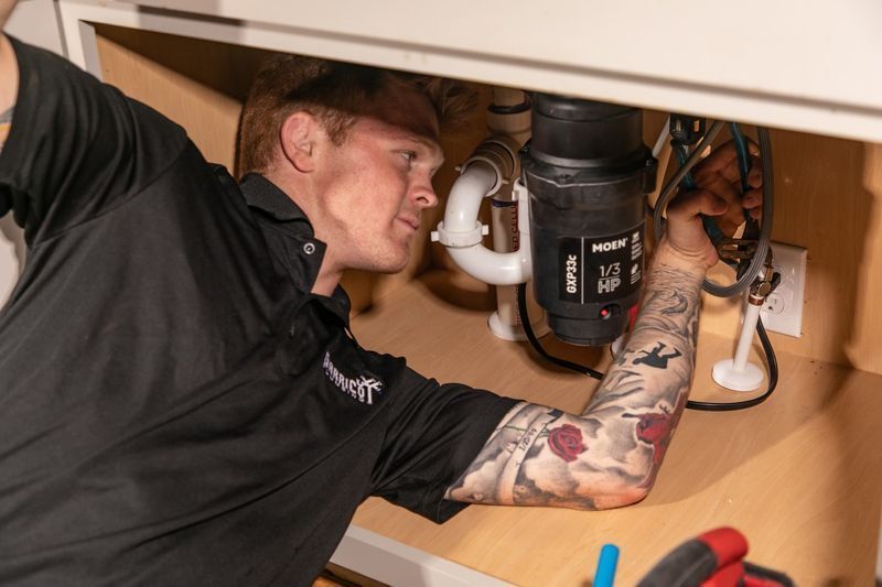 A plumber in a black shirt works on a garbage disposal unit under a kitchen sink.