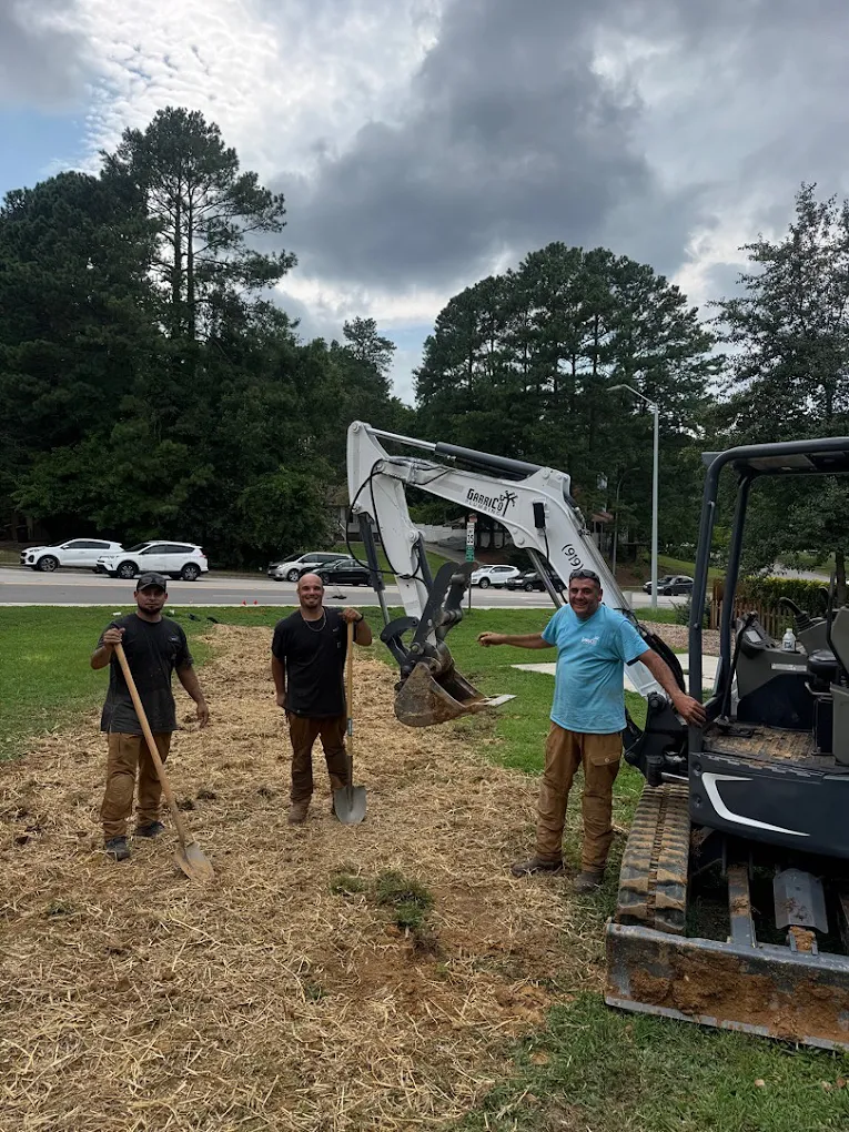 Three people stand in a field with wood chips, a shovel, and a white mini-excavator under a cloudy sky.