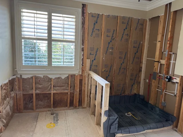 Bathroom under renovation, showing exposed wall studs, insulation, plumbing, and a shower base near a window.