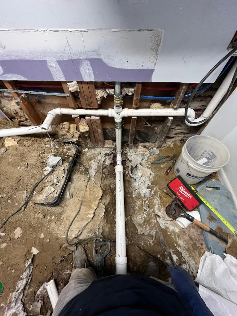 A view from above looking down at white PVC plumbing pipes installed in an open wall cavity and trench in the dirt floor.