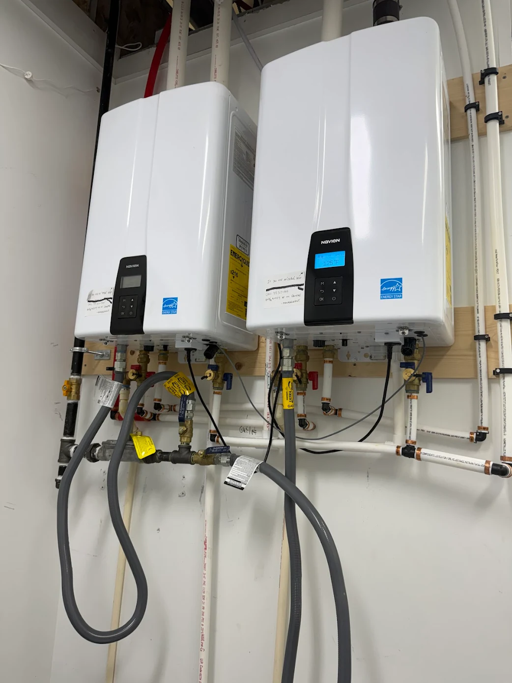 Two white tankless water heater units mounted side-by-side on a white wall with various connected pipes and valves.