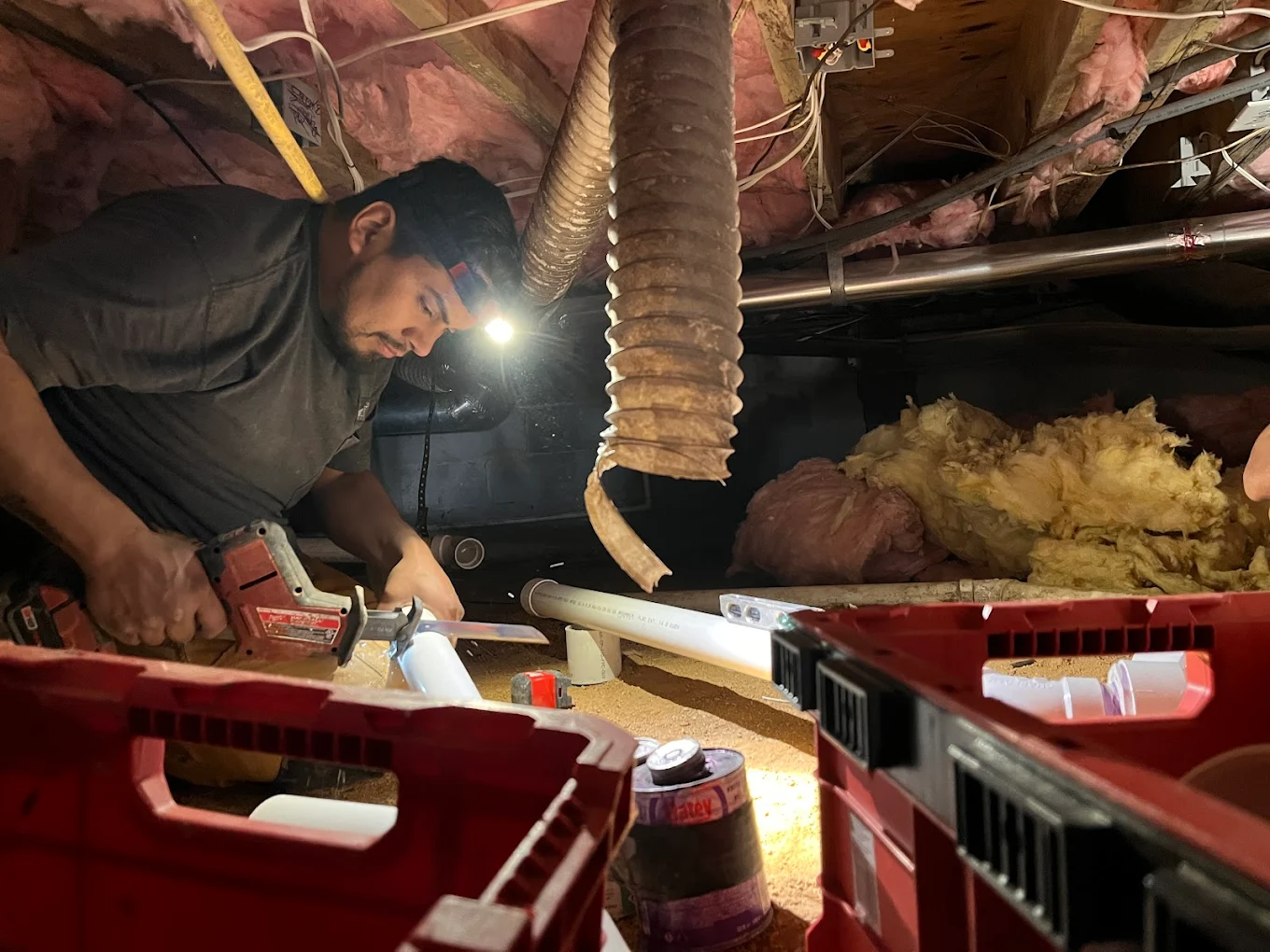 A person uses a reciprocating saw to cut a pipe in a cramped crawl space with exposed insulation and HVAC ducts.