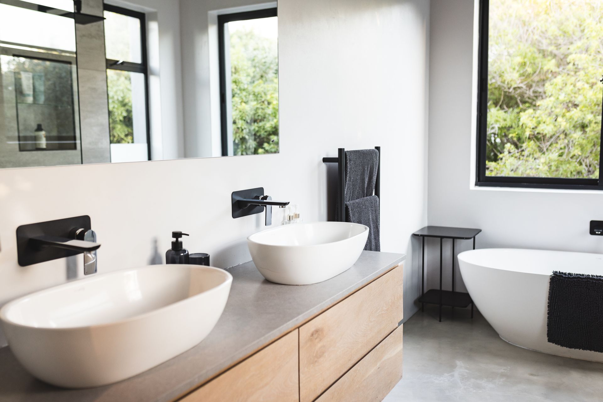Modern bathroom with double white vessel sinks, matte black wall-mounted faucets, a wooden vanity, and a freestanding tub.