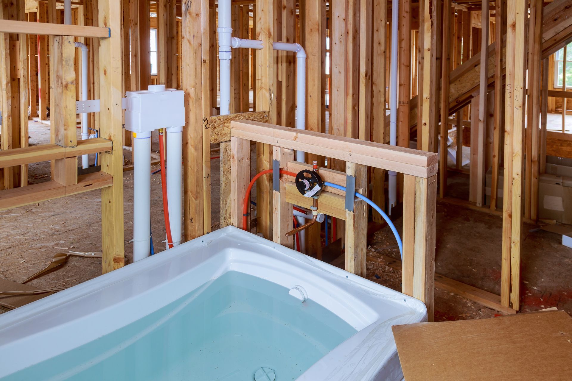 An unfinished room under construction showing a white bathtub, exposed wooden wall studs, and plumbing pipes and fixtures.