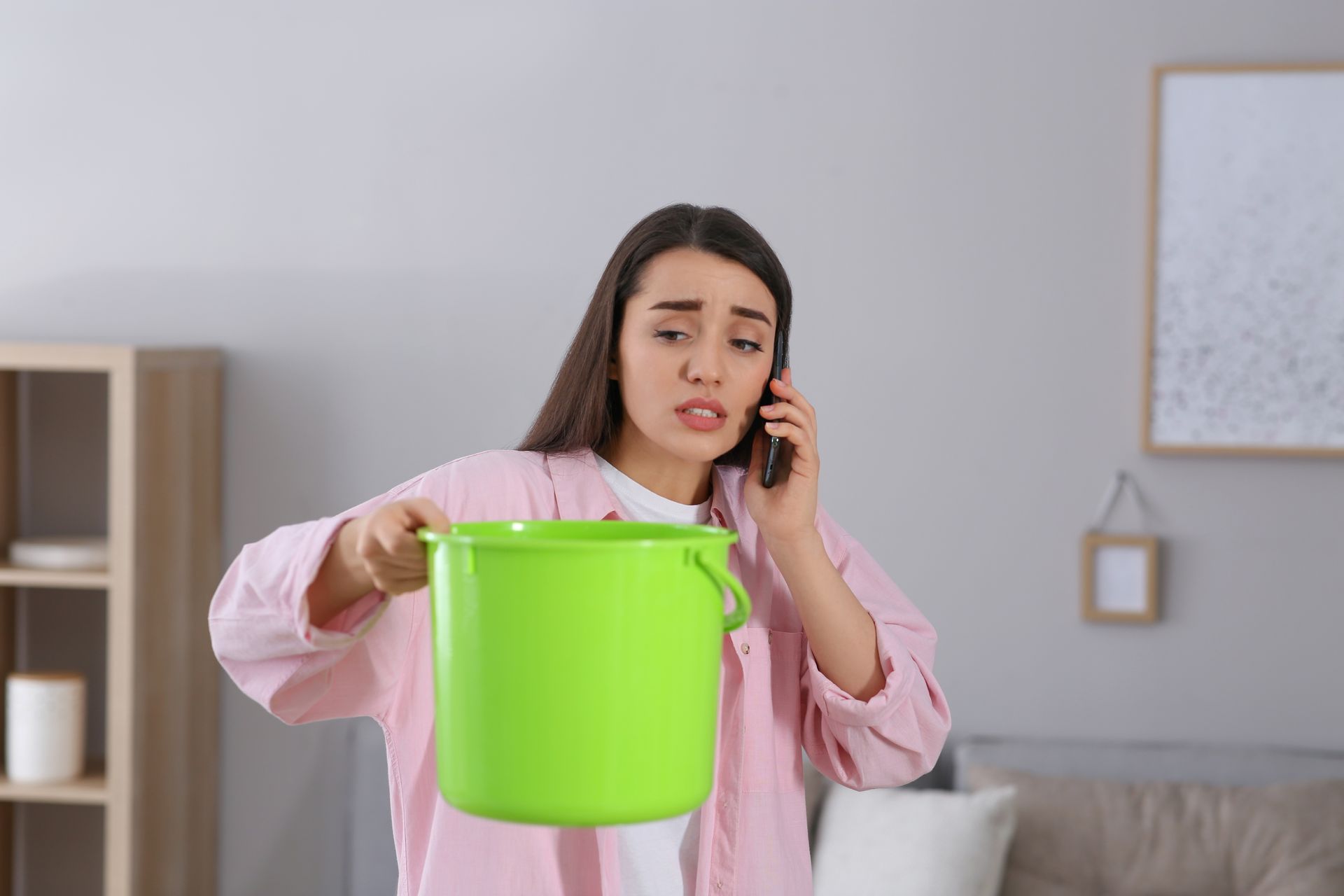 A person in a pink shirt looks concerned while holding a green bucket and talking on a phone in a living room.