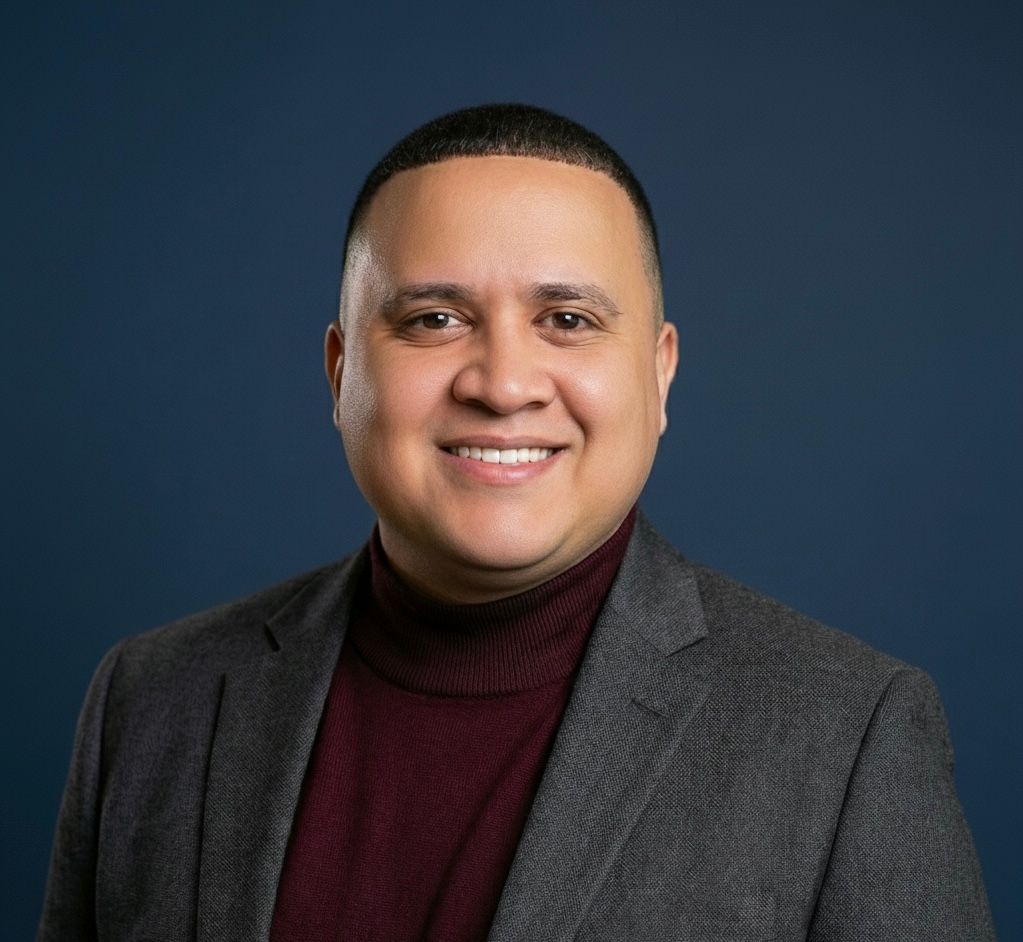 Man smiling, wearing a burgundy turtleneck, gray blazer. Blue background.
