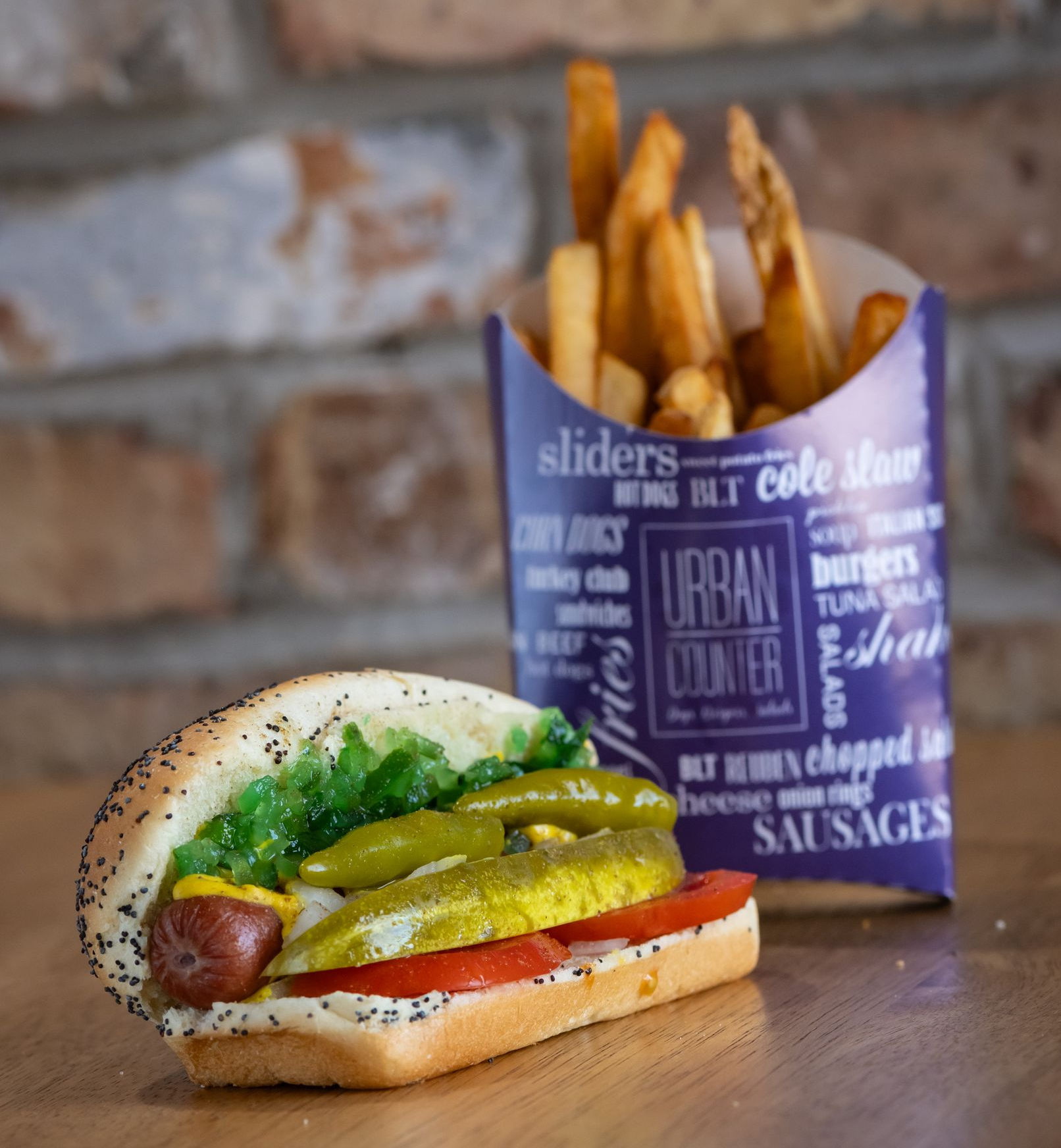 Chicago-style hot dog with toppings, next to a container of fries. Brick background.