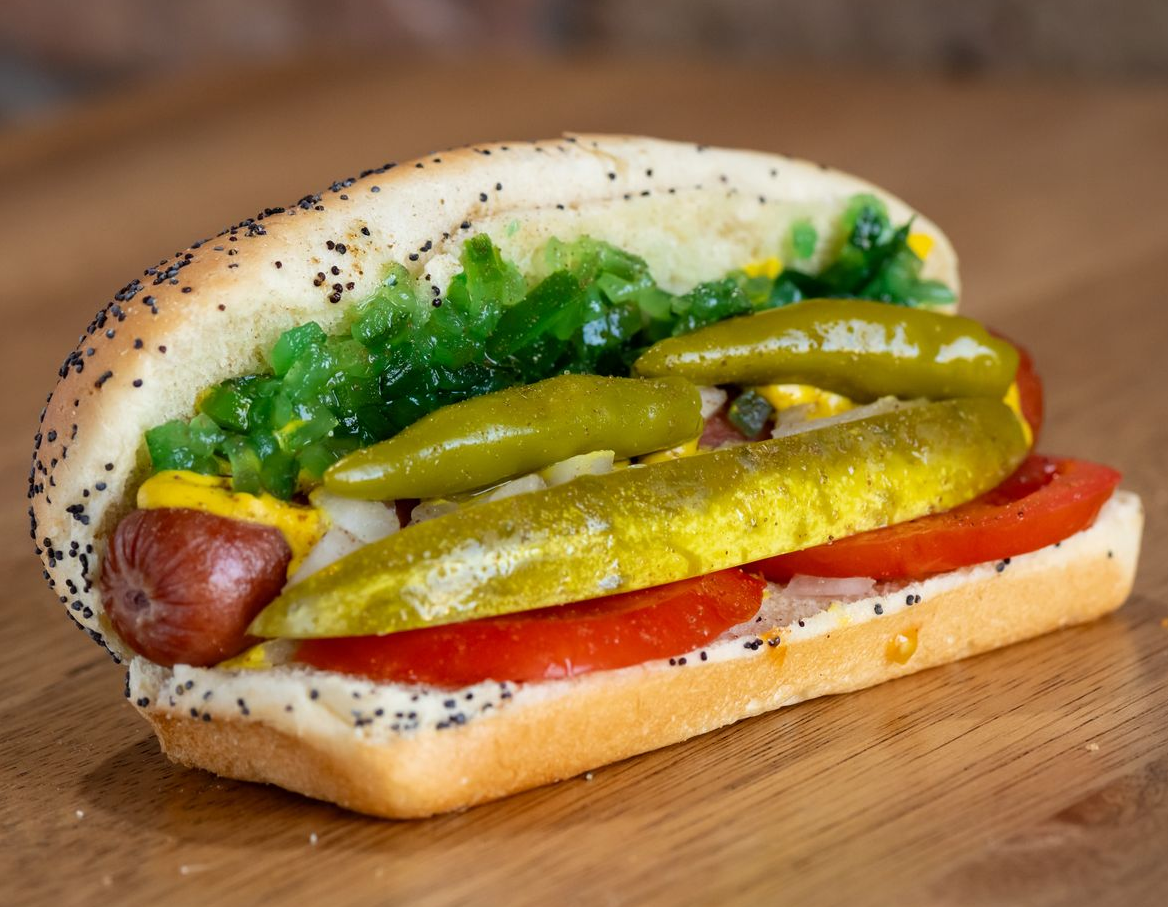 Close-up of a Chicago-style hot dog in a poppy seed bun, with toppings including pickle, tomato, and onion.