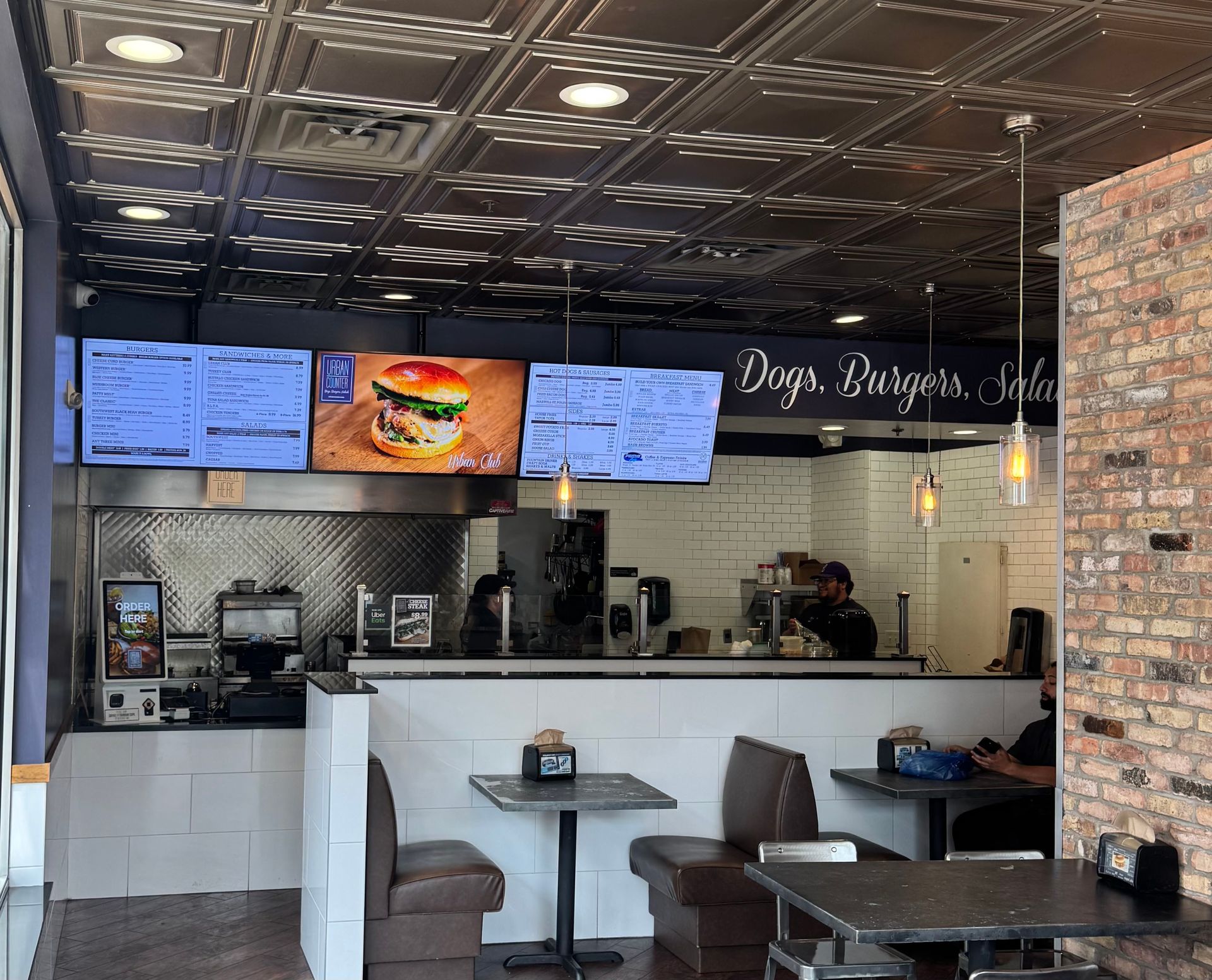 Interior of a restaurant: Counter with menus, tables, seating, and a brick wall.