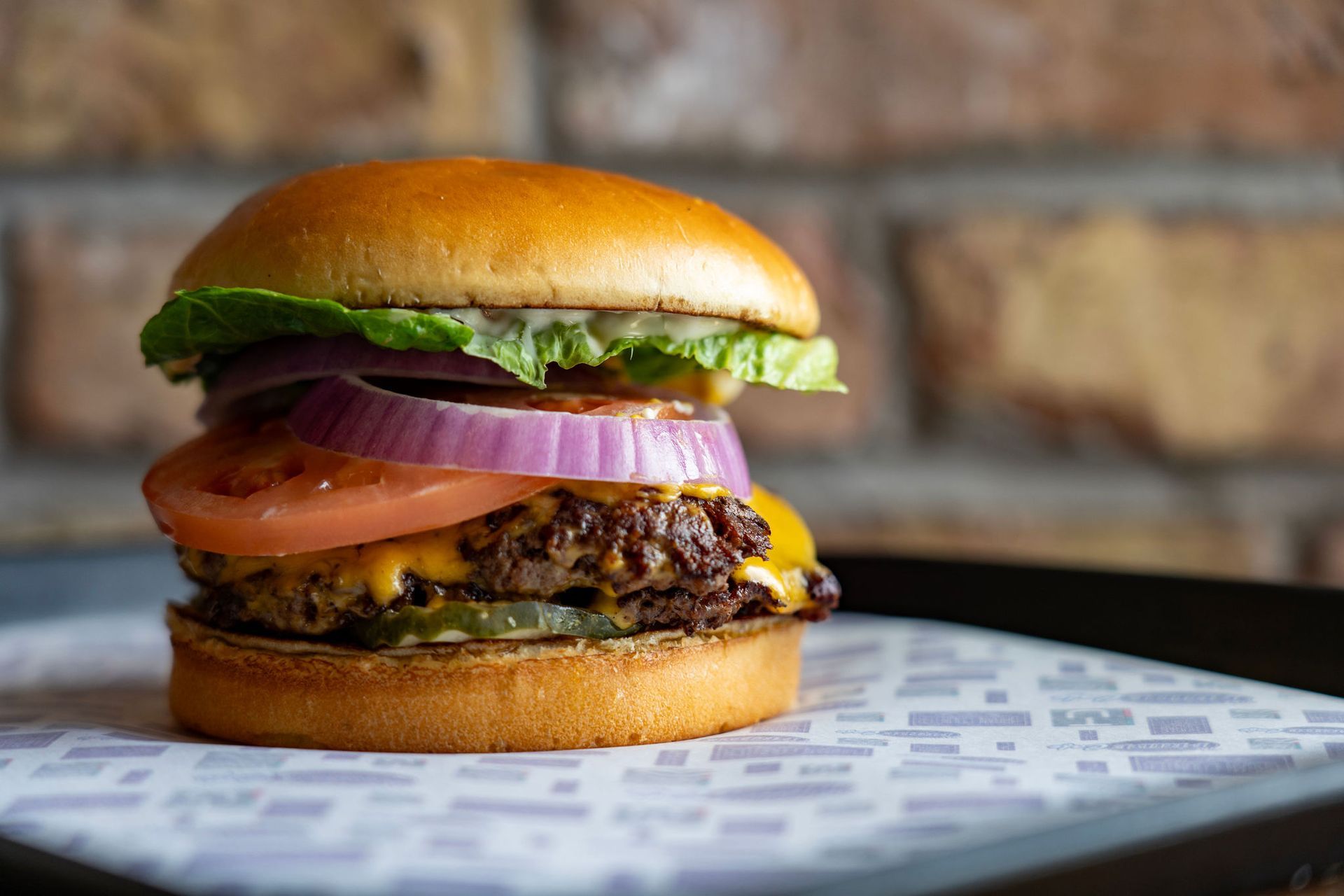 Cheeseburger on a bun with lettuce, tomato, red onion, and pickles on a patterned tray.