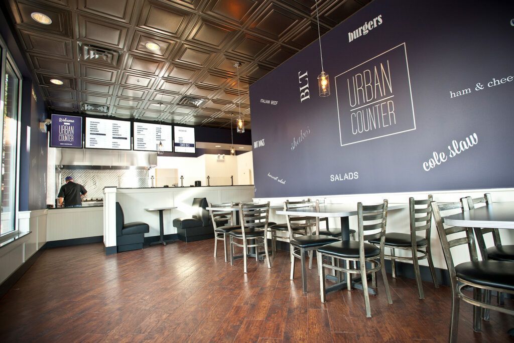 Interior of a restaurant with tables, chairs, and a large wall with menu and logo.