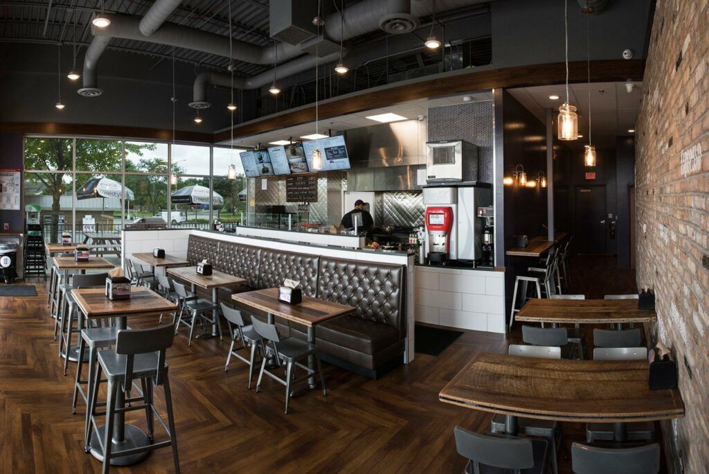 Restaurant interior: tables, stools, brick wall, open kitchen with workers, and large windows.