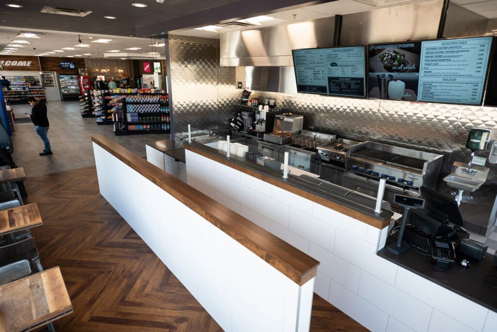 Inside a quick-serve restaurant, counter with stainless steel appliances, menu screens, and seating area with wooden tables.