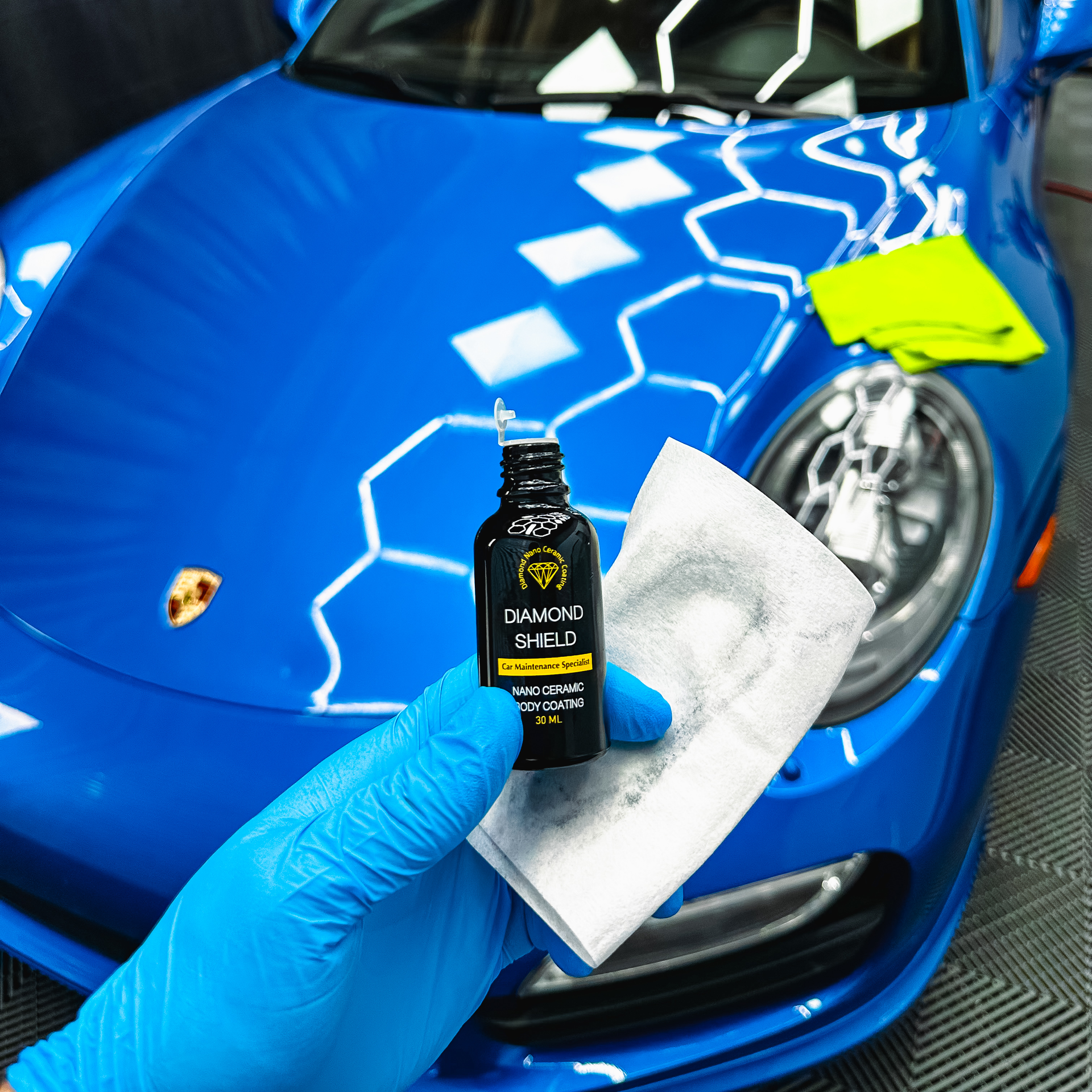 A gloved hand holds a bottle of car detailing product and a white applicator pad in front of a blue sports car.