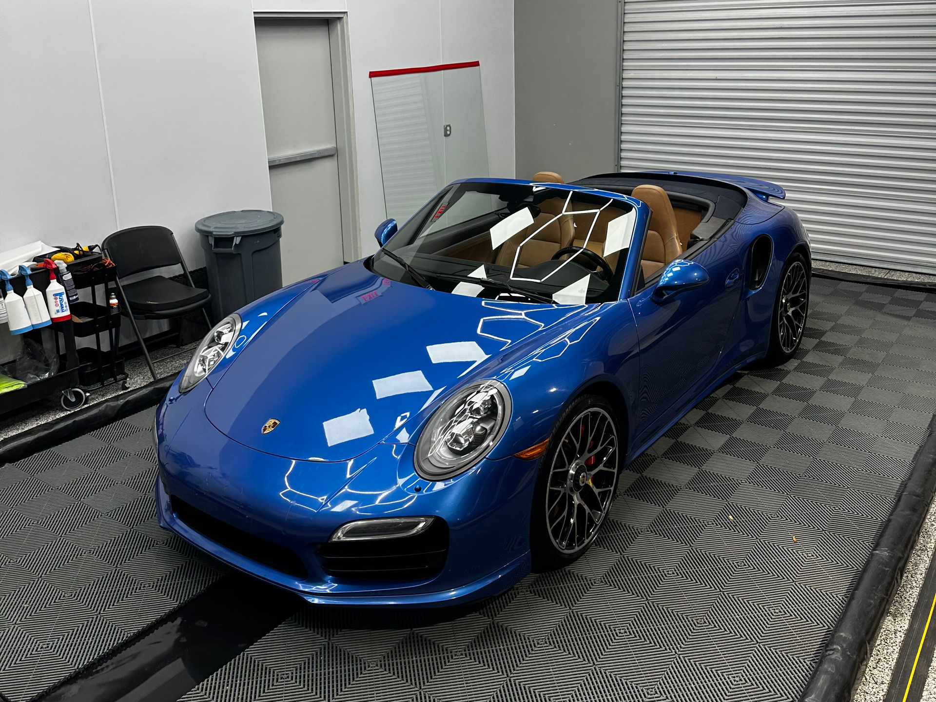 Blue Porsche convertible in a well-lit garage. Car sits on a patterned black floor, with detailing supplies nearby.
