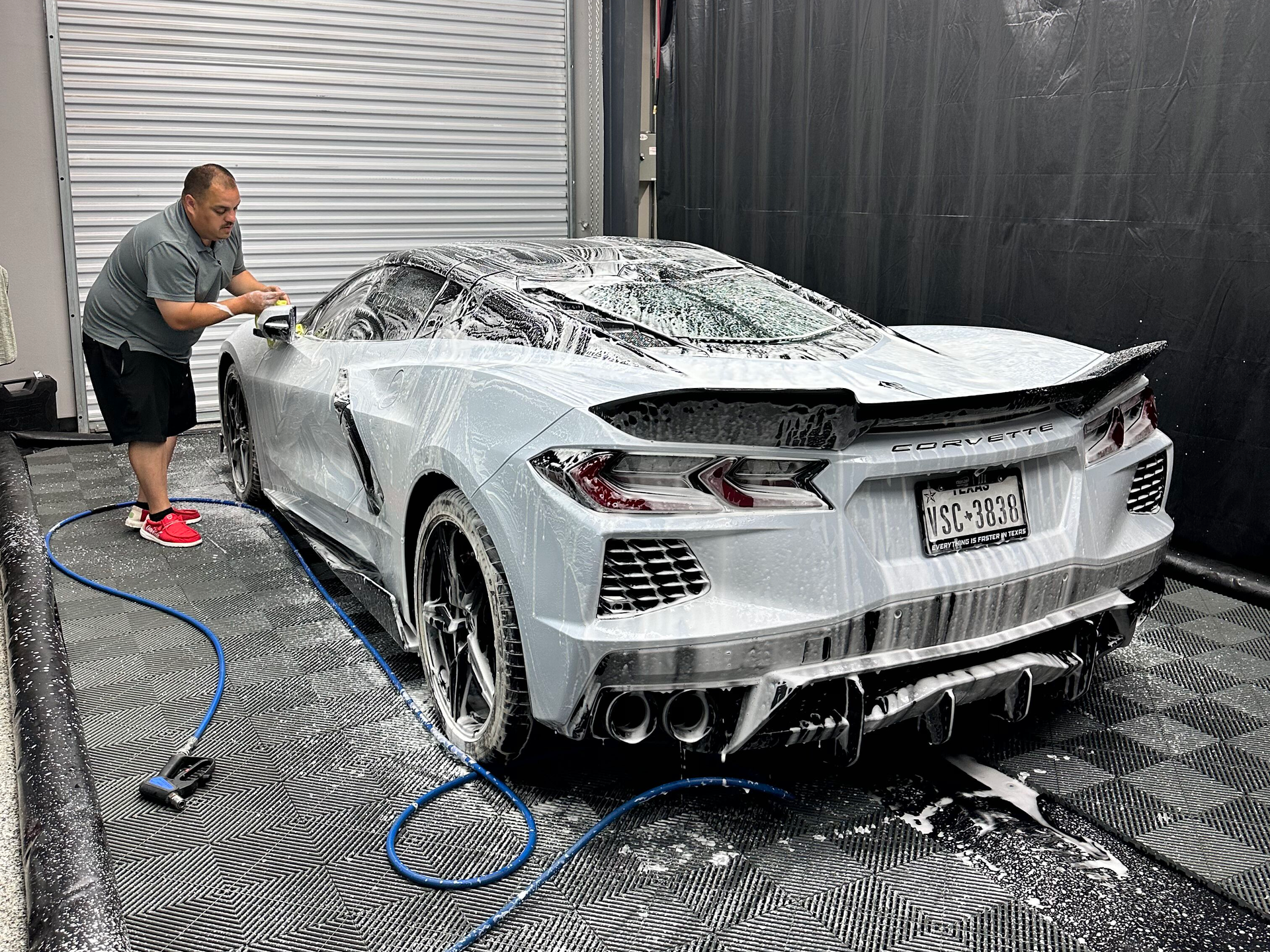 Man washing a light gray Corvette with white soap in a garage with black tile flooring and a gray wall.
