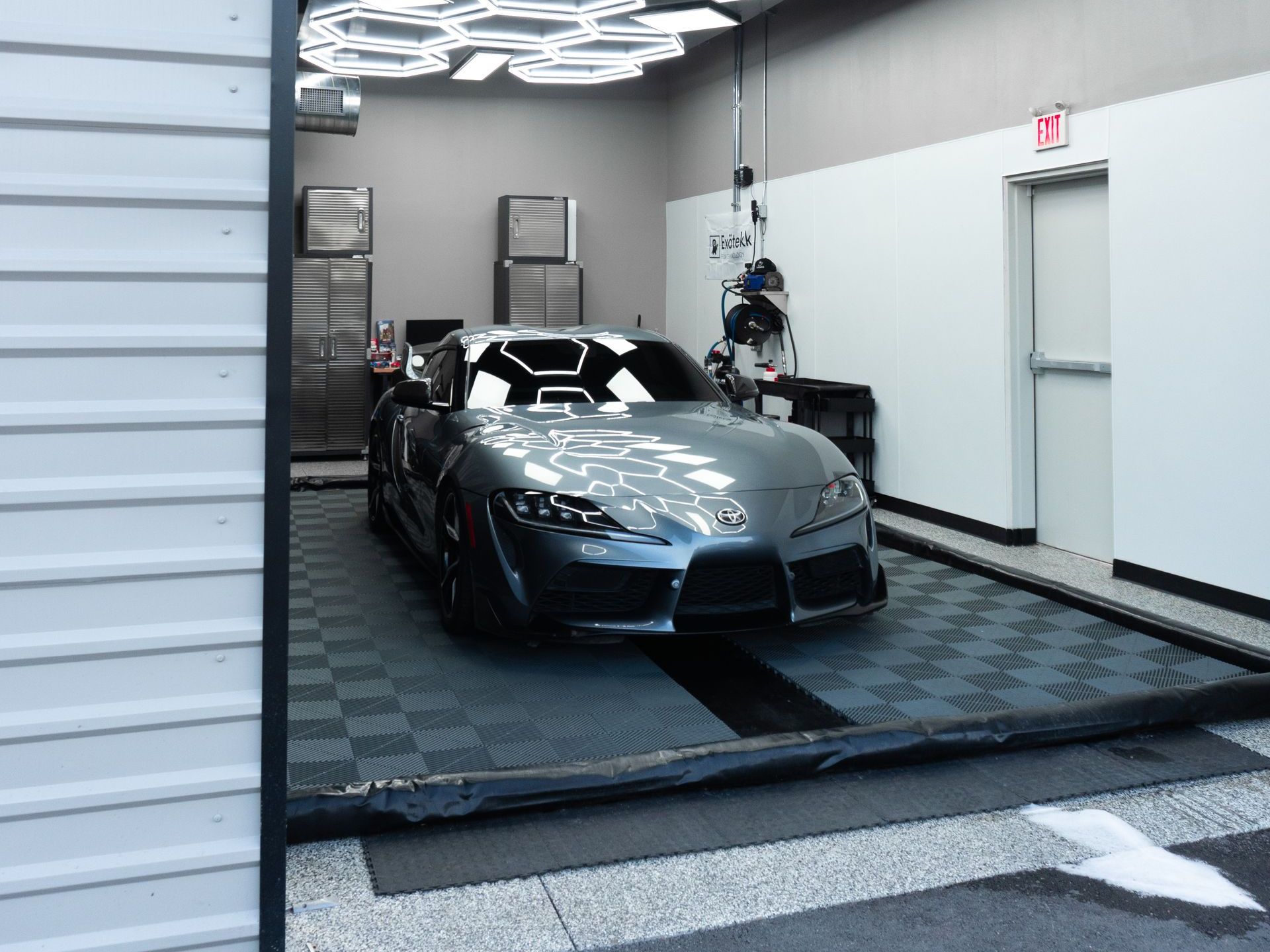 A gray sports car inside a garage with bright overhead lights. The car is parked on a tiled floor.