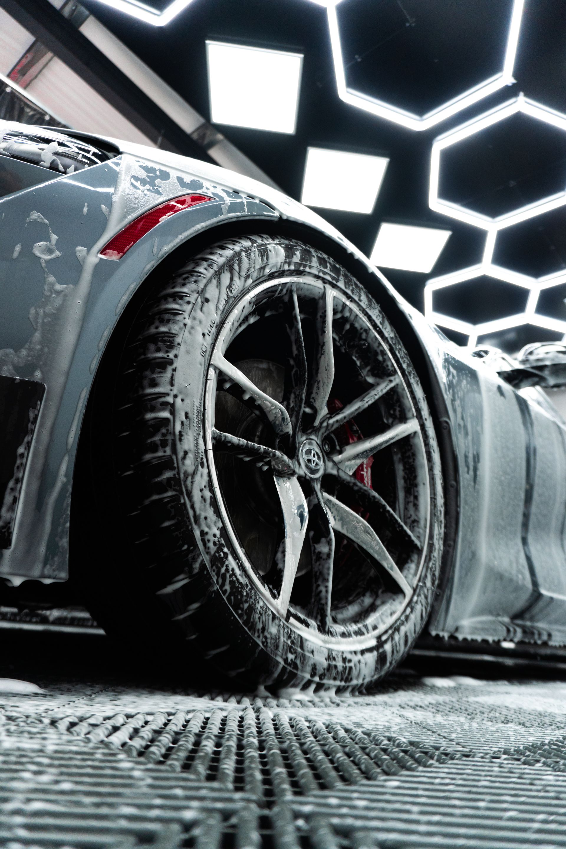 A car tire covered in soapy foam in a car wash. Overhead, hexagonal lights illuminate the scene.