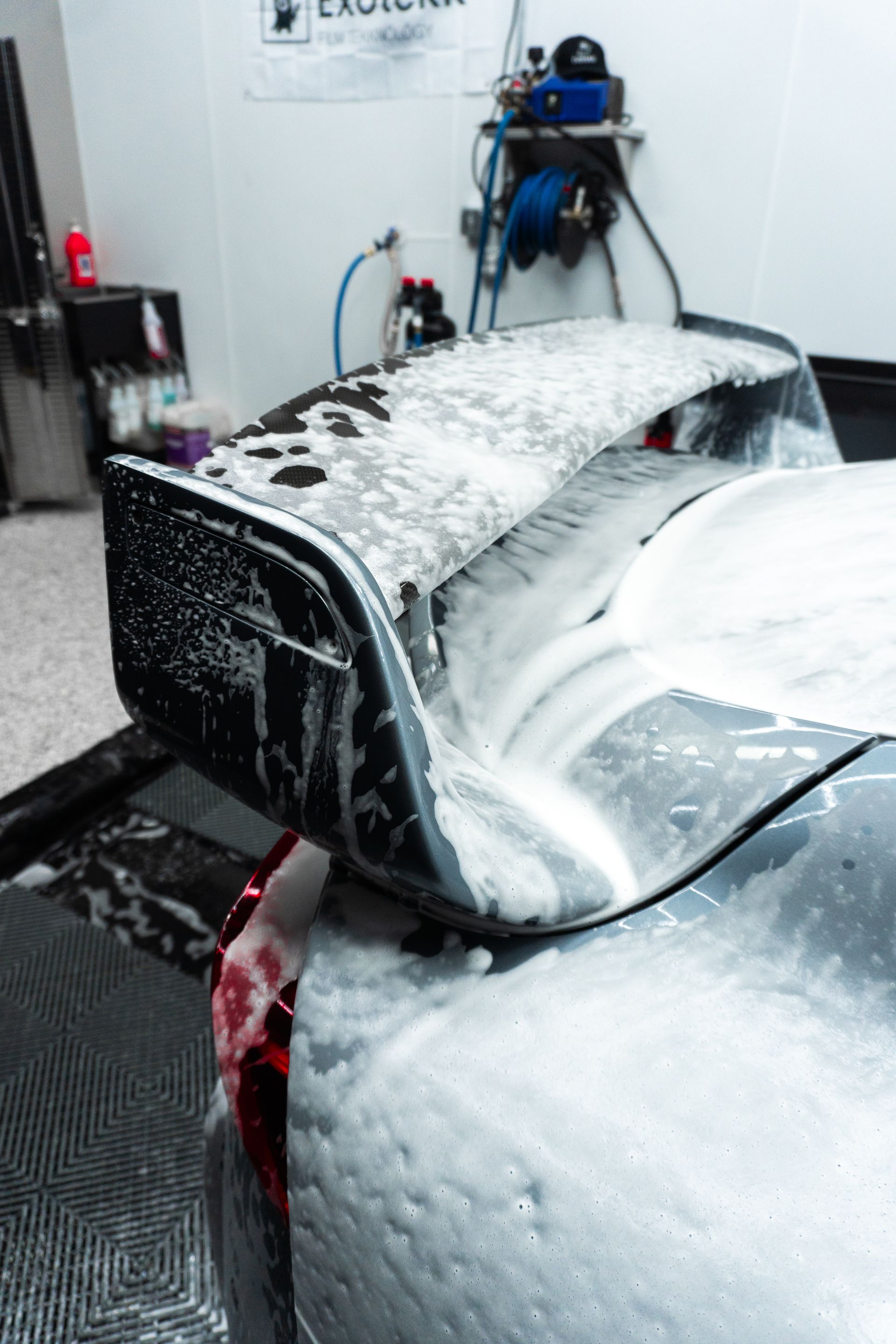 A car's rear wing covered in white foam, being washed at a car detailing shop.
