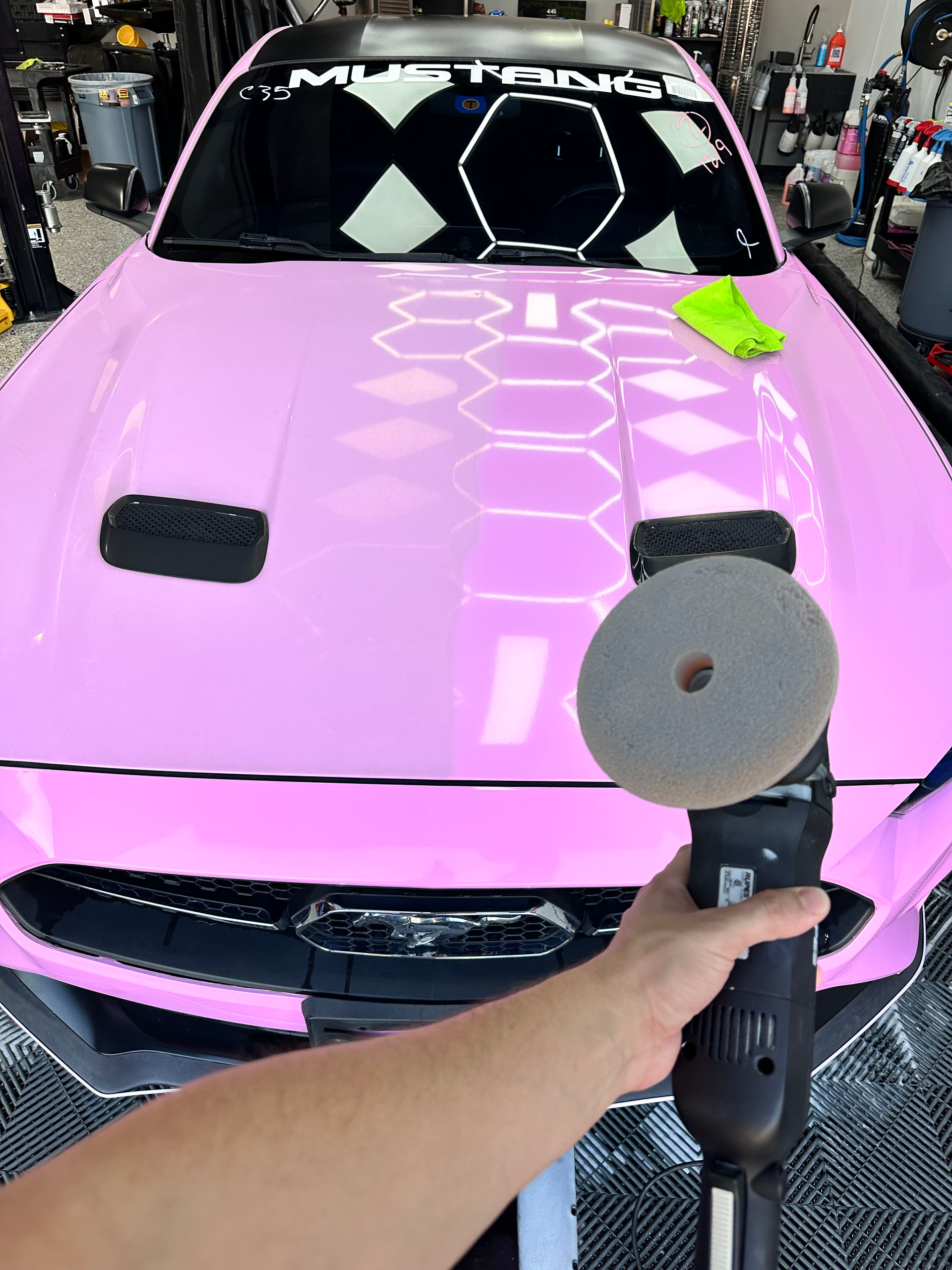 A person polishes a pink Mustang hood with a buffing machine. The hood features a honeycomb pattern and black vents.